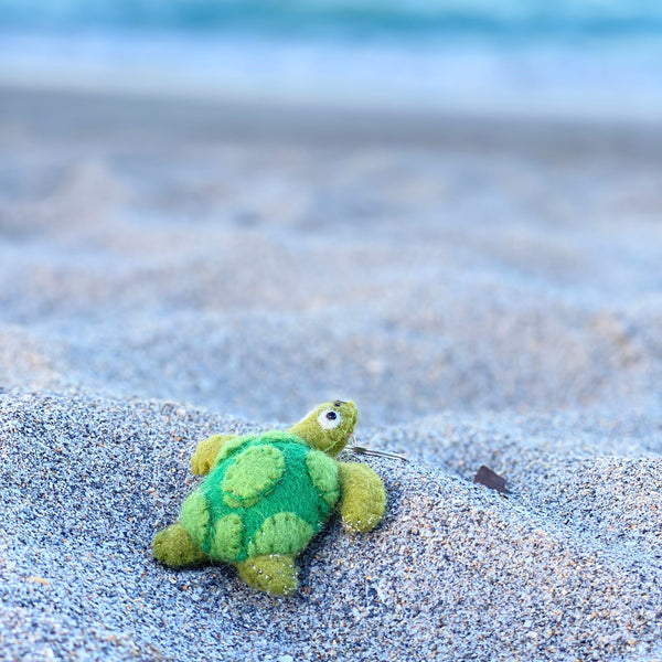 A green wool felt sea turtle keychain rests on a serene, sunlit sandy beach, with a blurred ocean in the background. This handmade felt keychain features a textured shell, small flippers, a head with a single visible eye, and a metal key ring attached.