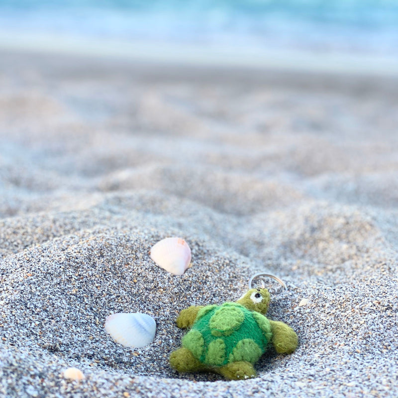 A handcrafted sea turtle felt keychain rests on grey sand with two seashells, set against a blurred beach and light blue ocean. This unique keychain features a mottled green wool shell with lighter edges, a darker pattern, fuzzy green legs, a head with a small black eye, and a metal keychain attached to its head.