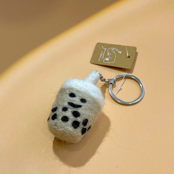 A handcrafted felt Taiwanese bubble tea keychain is displayed on a peach-colored surface, embodying a calm appeal. This unique keychain, shaped like a bubble tea drink, features a dark body with black bubble details and a white plush felt topping. It includes a metal keychain with a key ring, with a small tag visible behind it.