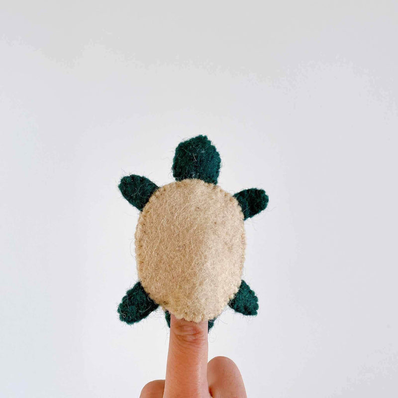 A charming needle felted turtle keychain is held up by a female hand against a plain white background. This unique wool felt turtle features a tan, oval-shaped body, a green felt head, and four matching green flippers, highlighting its delightful 3D design.