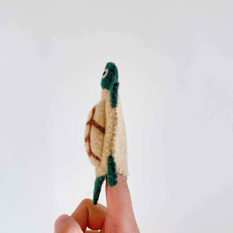 A handcrafted needle felted turtle keychain is presented in a female hand against a plain white backdrop. This charming felt keychain features a rounded green and tan turtle body with four green flippers, a green head, and brown stitching detailing its shell, showcasing its artisan quality.