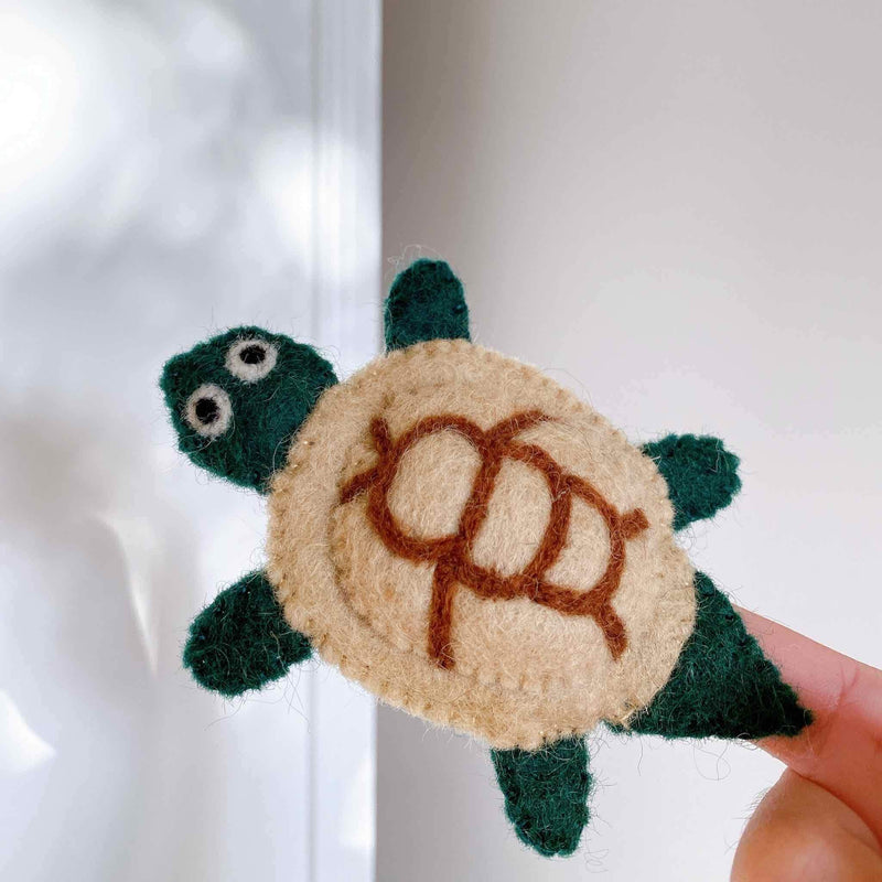 A wool felt turtle keychain is held in a female hand against a bright, soft-focused background. The 3D designed turtle features a dark green head, legs, and tail, with a tan shell patterned in brown. This needle felted, handcrafted keychain emphasizes its tactile and unique quality.