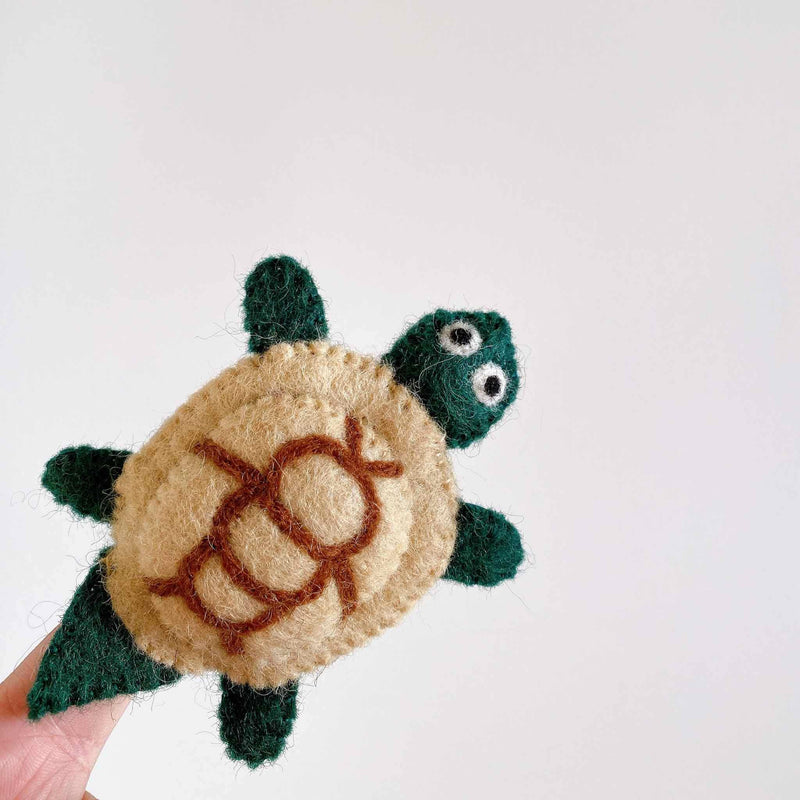 A charming wool felt turtle keychain is held by a female hand against a plain white background. The turtle has a tan wool felt shell with brown markings and darker green wool felt for its head and four flippers. White and black eyes complete its adorable look.