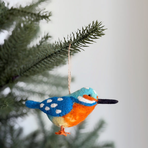 A vibrant wool felt Kingfisher ornament hangs on a Christmas tree branch, showcased in a cheerful close-up view. This unique Christmas ornament features vibrant blue wings with white speckles, an orange chest, and a distinctive black beak. A natural jute string is visible, used for hanging the handcrafted felt ornament.