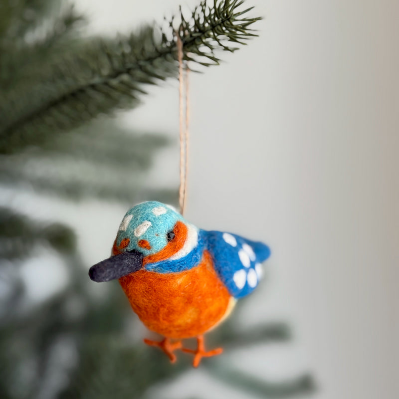 A handmade felt Kingfisher ornament hangs from a festive Christmas tree branch, presented against a soft white background. This detailed Kingfisher ornament features a light blue head speckled with white, a bright orange chest, and blue wings dotted with white. It has a sleek dark beak, tiny orange feet, and hangs gracefully by a jute string, adding a unique handcrafted charm.