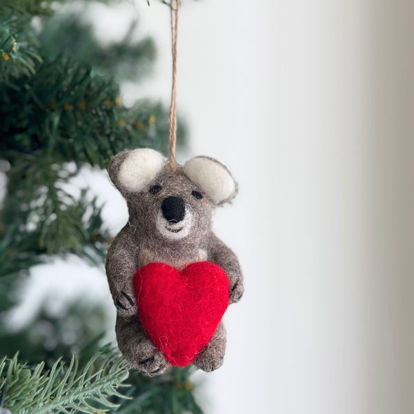 A heartwarming wool felt koala ornament, featuring a koala holding a heart, hangs on a Christmas tree. The gray koala has white inner ears and a black nose, sweetly clutching a bright red heart. A jute string loop at the top allows for easy hanging, evoking a sense of warmth and affection.