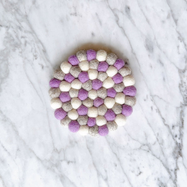 A single round lavender felt coaster, ethically handcrafted from 100% organic lamb wool felt balls, rests on a white marble background. This unique felt coaster features an evenly textured surface created by individually hand-stitched half-inch felt balls, measuring approximately 4 inches in diameter.