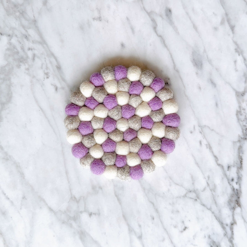 A single round lavender felt coaster, ethically handcrafted from 100% organic lamb wool felt balls, rests on a white marble background. This unique felt coaster features an evenly textured surface created by individually hand-stitched half-inch felt balls, measuring approximately 4 inches in diameter.