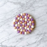 A handcrafted light pink and purple round felt coaster, part of a set of four, rests on a light marble surface. Made from 100% organic lamb wool, each approximately half-inch felt ball is hand-stitched to form its circular shape, highlighting its ethical craftsmanship. This unique felt coaster offers textural richness and charming, fair-trade warmth.