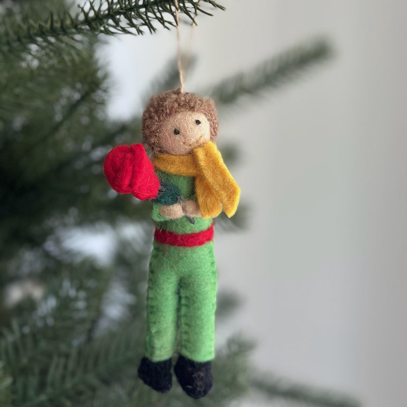 A handmade felt Little Prince ornament is suspended from a green Christmas tree branch against a soft white background. The charming felt ornament depicts the Little Prince with light brown hair, a yellow scarf, a green suit with a red belt, and black shoes, holding a red rose. It hangs from a natural jute string, evoking a calm and festive holiday feel.
