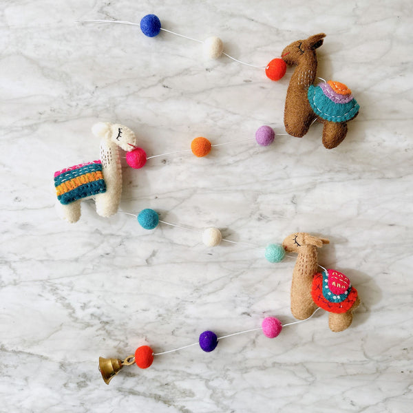 A playful wool felt llama garland is arranged on a marbled white surface. It features three 3D needle-felted llamas in shades of brown and white, each with a vibrant blue, orange, and pink striped saddle blanket and a small pink felt ball in its mouth. Colorful blue, orange, purple, and white felt balls are interspersed along the thin white string.