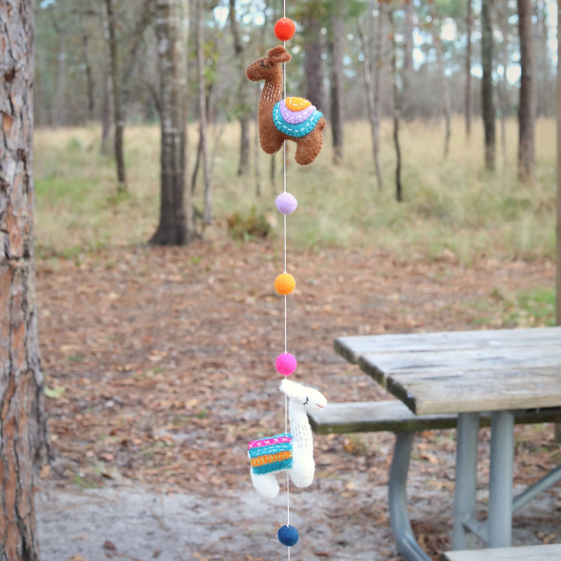 A wool felt Llama garland hangs playfully outdoors in front of a wooded area and a weathered picnic table. This handcrafted garland features two visible 3D felt llamas, one brown and one white, both adorned with colorful saddle blankets. Small, round felt balls in orange, pink, purple, and blue are interspersed along the string, adding a whimsical touch.