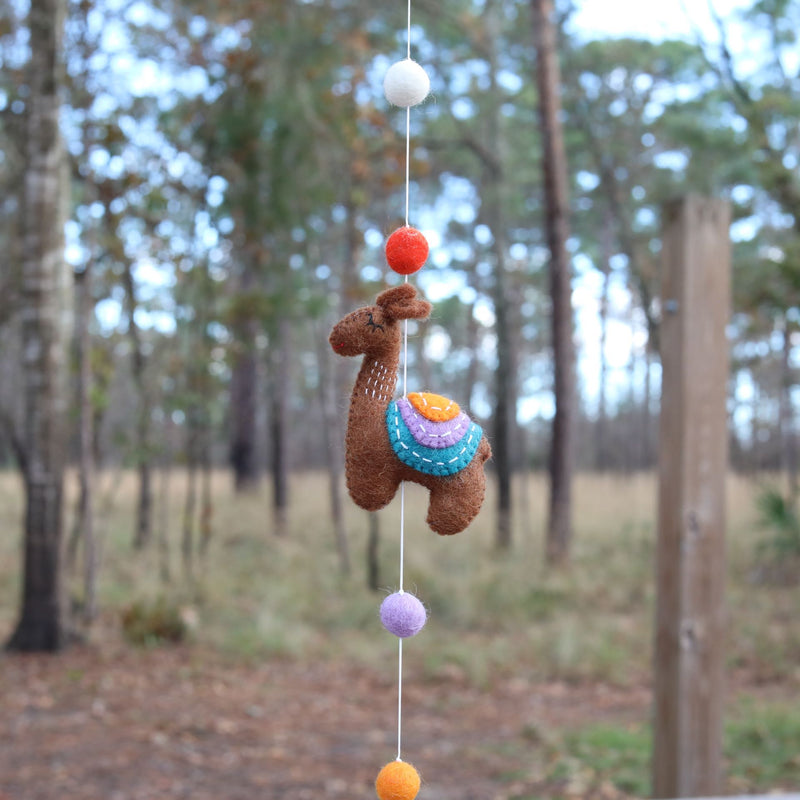 A close-up of a wool felt llama from a charming felt llama garland hangs by a white string in an outdoor forest. This skillfully needle-felted brown llama features a multi-colored saddle with vibrant orange, purple, and teal layers, enhanced by white stitch details. Colorful felt balls are interspersed along the string, adding to the garland's festive charm.