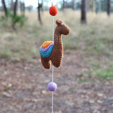 A whimsical brown wool felt llama, part of a felt llama garland, hangs playfully against an out-of-focus backdrop of trees and foliage. The centered llama features a colorful saddle with stitched orange, lavender, and teal layers. It is suspended by a string connected to an orange felt ball above and a purple felt ball below, highlighting its handcrafted and playful design.