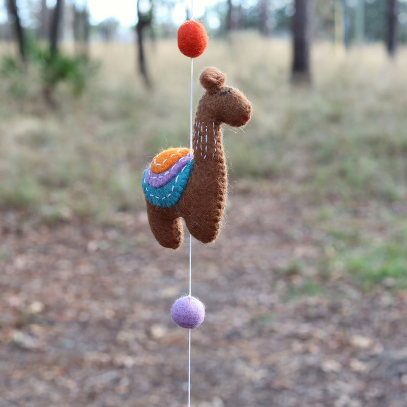 A whimsical brown wool felt llama, part of a felt llama garland, hangs playfully against an out-of-focus backdrop of trees and foliage. The centered llama features a colorful saddle with stitched orange, lavender, and teal layers. It is suspended by a string connected to an orange felt ball above and a purple felt ball below, highlighting its handcrafted and playful design.