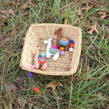 A charming wool felt llama garland is displayed in a light brown woven basket nestled in vibrant green grass. This whimsical garland features three skillfully needle-felted 3D llamas: one light brown, one white, and one tan. Each llama has a colorful saddle blanket with bright orange, turquoise, pink, and white stitching. Colorful orange, purple, and white felt balls accent the garland's white cord.