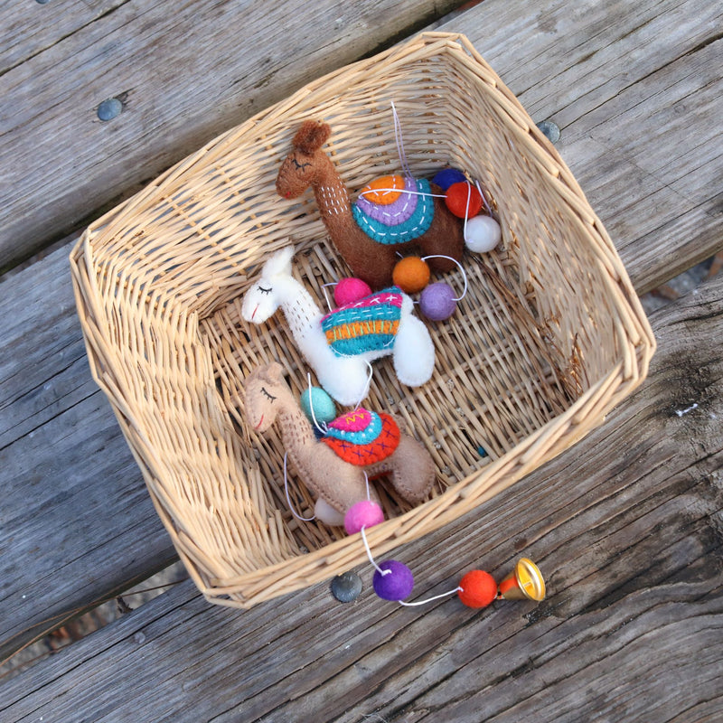 A cheerful wool felt llama garland is displayed inside a light brown woven basket, resting on a weathered wooden surface. This charming handmade felt garland features three felt llamas, each with small stitched eyes and a cheerful expression. The llamas are colored light brown, white, and tan, each adorned with a multi-colored saddle, strung alongside colorful felt balls.
