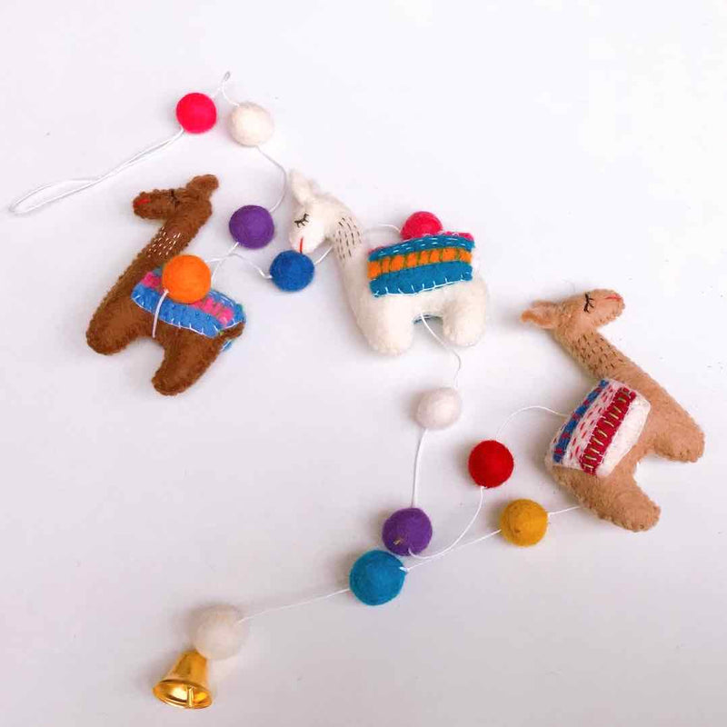 A playful wool felt llama garland is displayed against a clean white background. This approximately 4-foot-long garland features three three-dimensional, needle-felted llamas in brown and white, each wearing vibrant blankets. Colorful felt balls in red, white, purple, blue, and yellow are strung along the white cord, adding to its festive charm.