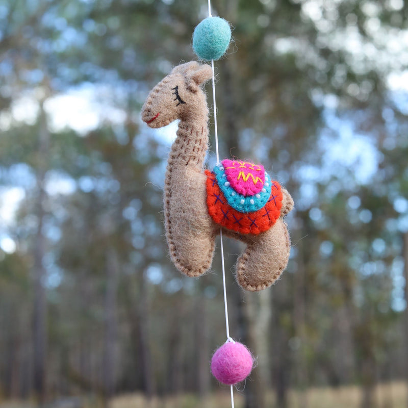 A handmade felt llama garland is featured, with a single light brown wool felt llama suspended by a white string against a softly blurred forest backdrop. This needle-felted llama showcases a captivating 3D design and wears a vibrant saddle with a pink, yellow, and blue geometric pattern. A teal felt ball hangs above it on the string, with a pink felt ball below, adding a whimsical touch.