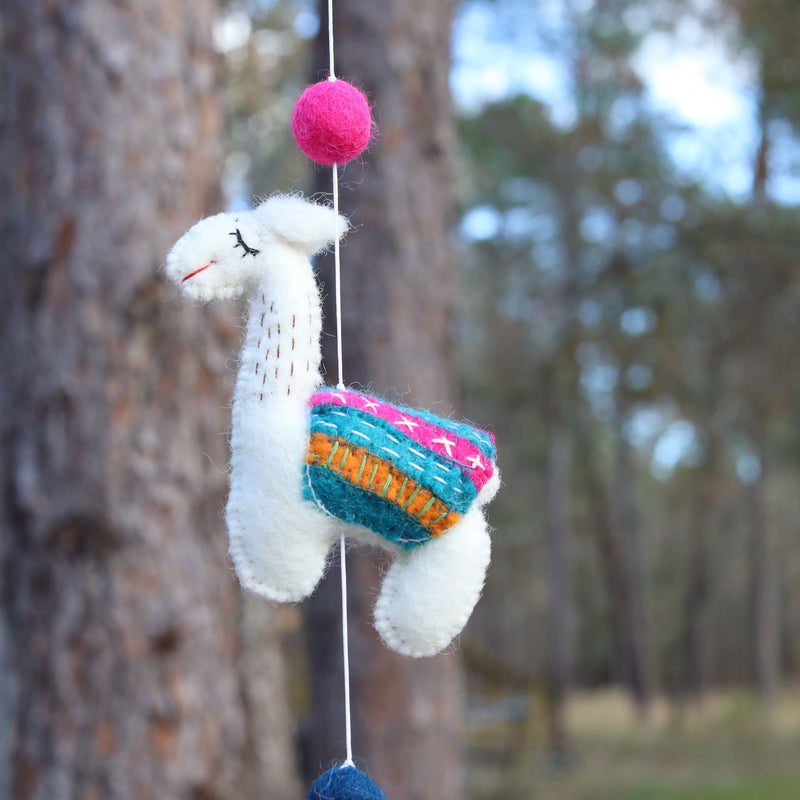 A charming white wool felt llama, part of a felt llama garland, is suspended outdoors against a blurred backdrop of trees and foliage. The handcrafted llama features a red stitched smile, black closed eye, and wears a vibrant blanket with teal, orange, and pink stripes accented by white stitches. It hangs from a white string between a pink ball above and a dark blue ball below, creating a whimsical and playful ambiance.