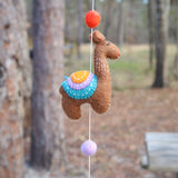 One brown wool felt llama from a felt llama garland is displayed outdoors against a blurred background of trees and leaves. The needle-felted llama features a decorative saddle in orange, lavender, and teal. An orange felt ball hangs above the llama, with a lavender felt ball below it, both attached by a white string.