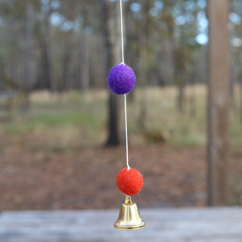 A close-up of a colorful wool felt ball detail from a Felt Llama Garland hangs outdoors against a blurred wooded background, creating a calm atmosphere. The round purple felt ball is suspended by a white string, with a second white string connecting to an orange felt ball below it.