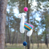 A single handcrafted wool felt llama from a felt garland is suspended against a softly blurred forest background, creating a whimsical feel. This white, needle-felted 3D llama features black stitched eyes, a red smile, and a colorful pink, orange, and teal saddle blanket. A bright pink felt ball is attached to the string above the llama, with a navy blue felt ball hanging below it.