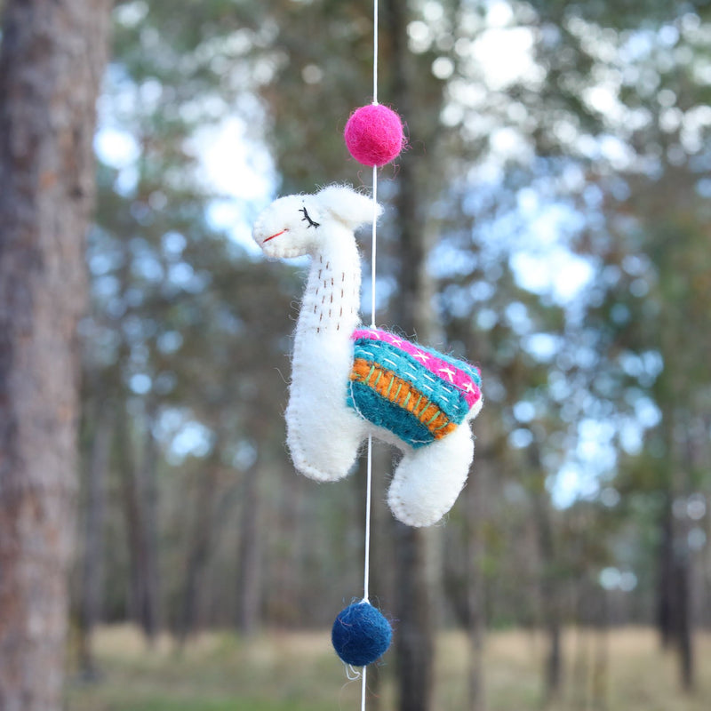A single handcrafted wool felt llama from a felt garland is suspended against a softly blurred forest background, creating a whimsical feel. This white, needle-felted 3D llama features black stitched eyes, a red smile, and a colorful pink, orange, and teal saddle blanket. A bright pink felt ball is attached to the string above the llama, with a navy blue felt ball hanging below it.