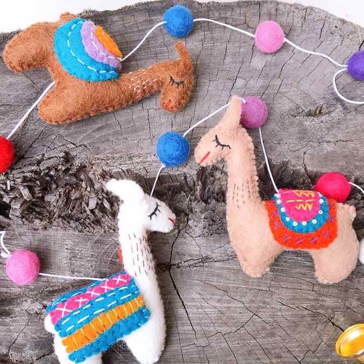 The felt llama garland is displayed on a rustic wooden surface, creating a whimsical scene. This handcrafted wool felt garland features three distinct llamas interspersed with colorful felt balls. One llama is light brown with an orange, blue, and purple saddle; another is light pink with a red, pink, and blue saddle; and the third is white with a pink, blue, and orange saddle. Red, pink, purple, and blue felt balls complete the vibrant design.