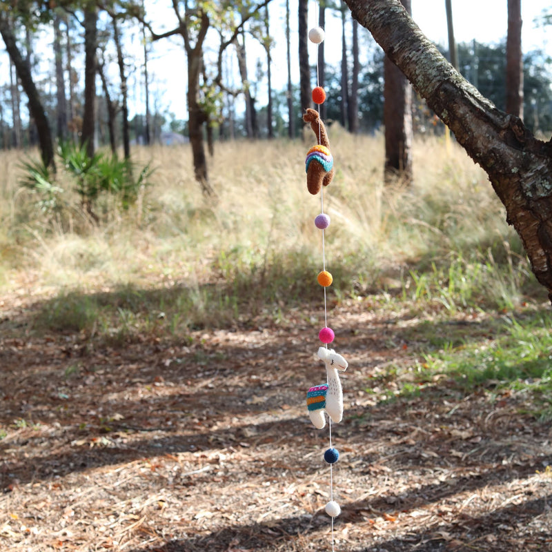 A colorful wool felt llama garland hangs gracefully from a tree branch, outdoors in a natural setting with tall grasses and trees in the background. The handcrafted garland features two distinct felt llamas, one brown and one white, both adorned with vibrant saddle blankets. Colorful felt balls in shades of white, purple, orange, and pink separate the playful llamas, creating a whimsical and festive mood.
