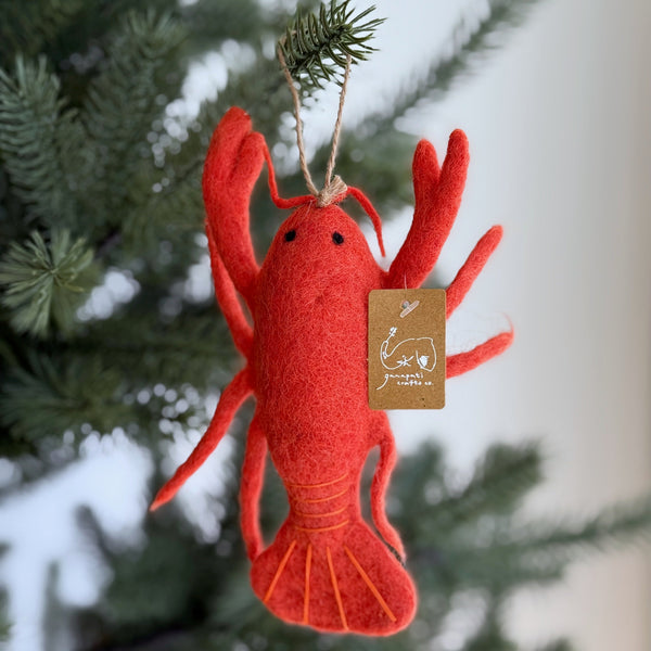 A vibrant red wool felt lobster ornament hangs on the green needles of a Christmas tree. This unique Christmas ornament is crafted with detailed claws, antennae, and a segmented tail, featuring a jute string for hanging. It adds a joyful, nautical charm to the holiday tree.