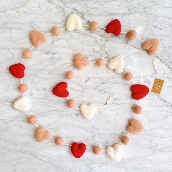 The felt love garland, made of felted wool, is displayed on a white marble surface, creating a cheerful decorative atmosphere. It features alternating red, blush pink, and white hearts with blush pink balls, all evenly spaced along the strand.