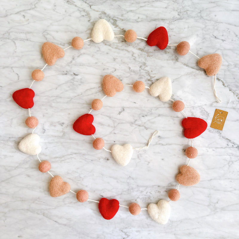 The felt love garland, made of felted wool, is displayed on a white marble surface, creating a cheerful decorative atmosphere. It features alternating red, blush pink, and white hearts with blush pink balls, all evenly spaced along the strand.