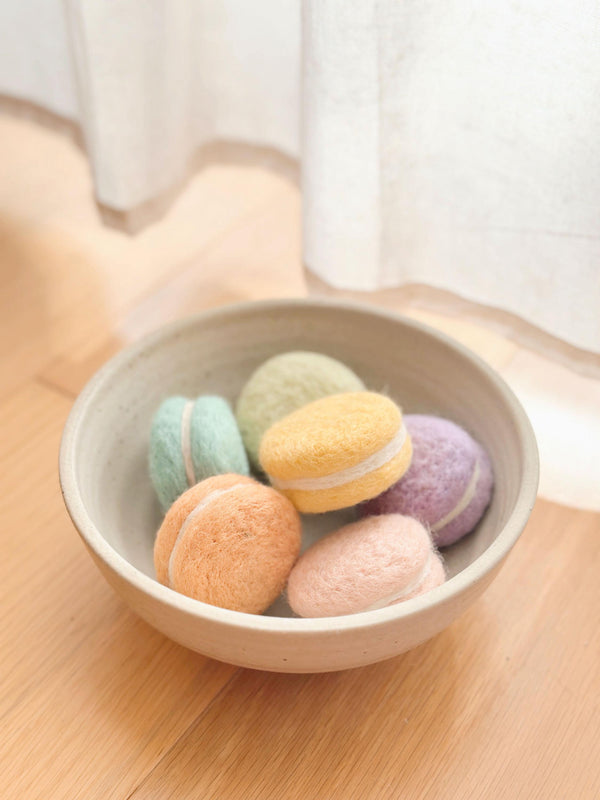 A delightful set of handcrafted wool felt macarons are artfully arranged in a light beige ceramic bowl, resting on a light brown wooden surface against a soft white curtain. These round, textured felt play food desserts come in pastel orange, pink, yellow, green, blue, and purple, each with a white filling. They are handmade with azo-free dyes, reflecting fine craftsmanship.