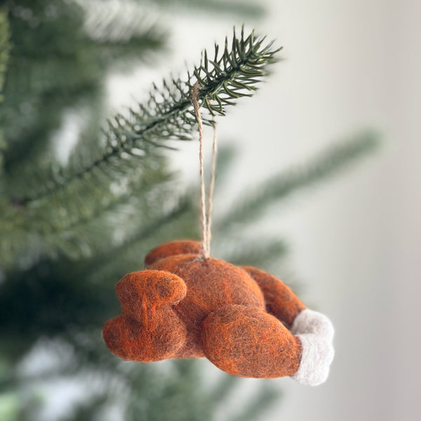 A handcrafted wool felt turkey ornament is displayed on a pine tree branch, evoking a warm and festive mood. This charming brown turkey features fluffy white details at its feet and a jute string at the top for easy hanging.
