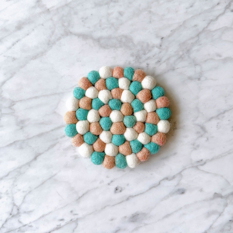 A round, handmade Felt Miami Round Coaster, crafted from 100% organic lamb wool, rests on a white marble surface. This unique wool felt coaster is composed of tightly clustered, hand-stitched felt balls in pastel shades of turquoise, peach, and white. Its eco-friendly design highlights the craftsmanship of Nepalese female artisans, adding a vibrant and tactile accent.
