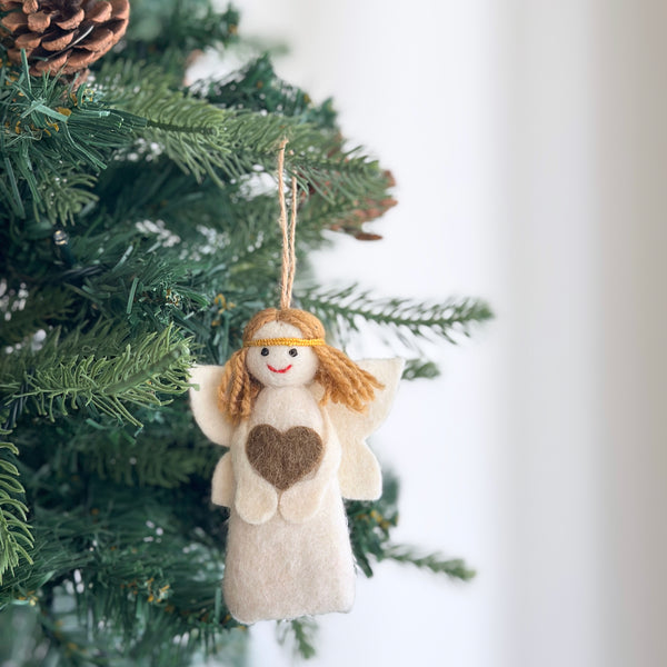 An off-white felt angel Christmas ornament hangs gracefully from a Christmas tree adorned with pine cones. This mini angel features a soft felt body and wings, a prominent brown heart on its chest, golden yarn hair with a golden band, and a red stitched smile, ready to be displayed by a jute string.