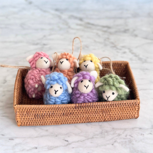 Six delightful pastel wool felt sheep ornaments are nestled in a woven rattan basket on a marbled surface. Each of these handmade felt ornaments, from a set of six, features a soft, fluffy texture and small white ears. The wool felt sheep ornaments come in charming pastel tones of pink, beige, yellow, blue, purple, and green, each with a jute string for hanging.