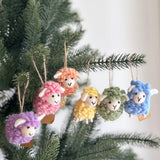 Six felt mini fluffy sheep ornaments are joyfully displayed hanging from the branches of a Christmas tree. Each wool felt sheep ornament is crafted in a unique pastel color, including purple, pink, beige, yellow, green, and blue. They feature a fluffy texture, small white ears, and simple stitched faces with a black 'V' mouth, adding a playful touch. A brown jute string suspends each charming ornament.