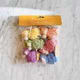 A set of six wool felt mini fluffy sheep ornaments, in pastel pink, beige, yellow, green, blue, and purple, are arranged in their transparent packaging on a white marble surface. Each felt sheep ornament has a fluffy texture and a jute string for hanging, perfect as a playful Christmas tree ornament.