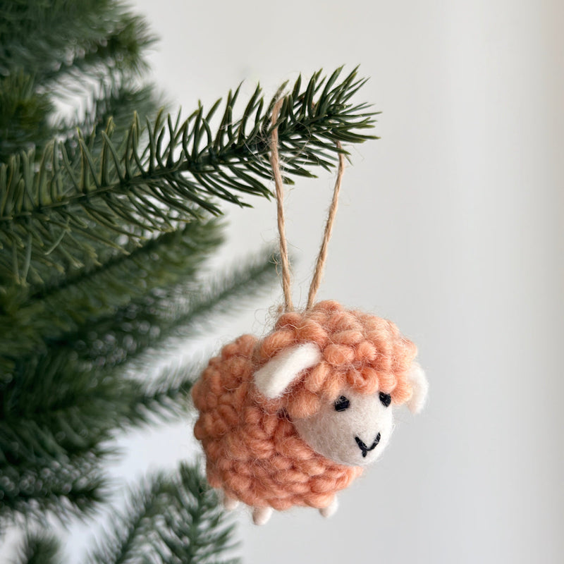 A peach-colored felt mini fluffy sheep ornament hangs on a green Christmas tree branch, set against a plain white wall. This playful handmade felt ornament features a fluffy body, small white felt ears and legs, black eyes, and a charming stitched black smile.