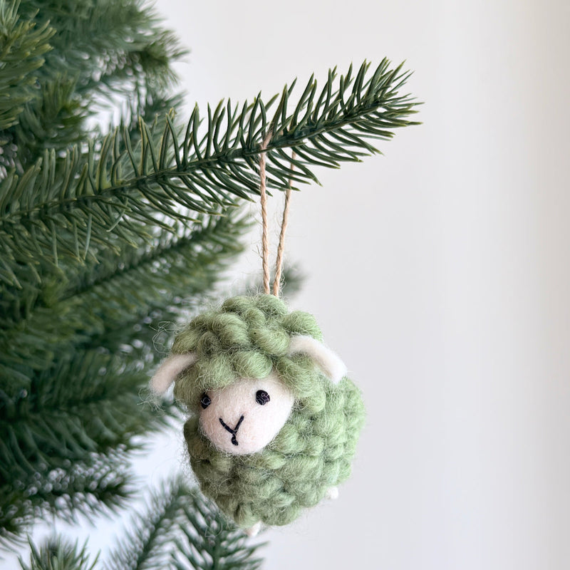 A pastel green felt mini fluffy sheep ornament hangs gracefully from a Christmas tree branch against a plain white background. This handmade felted wool sheep ornament features a textured pastel green body, small white ears, and a white felt face with simple black eyes and a stitched 'Y' mouth. A jute string loop is attached for hanging.