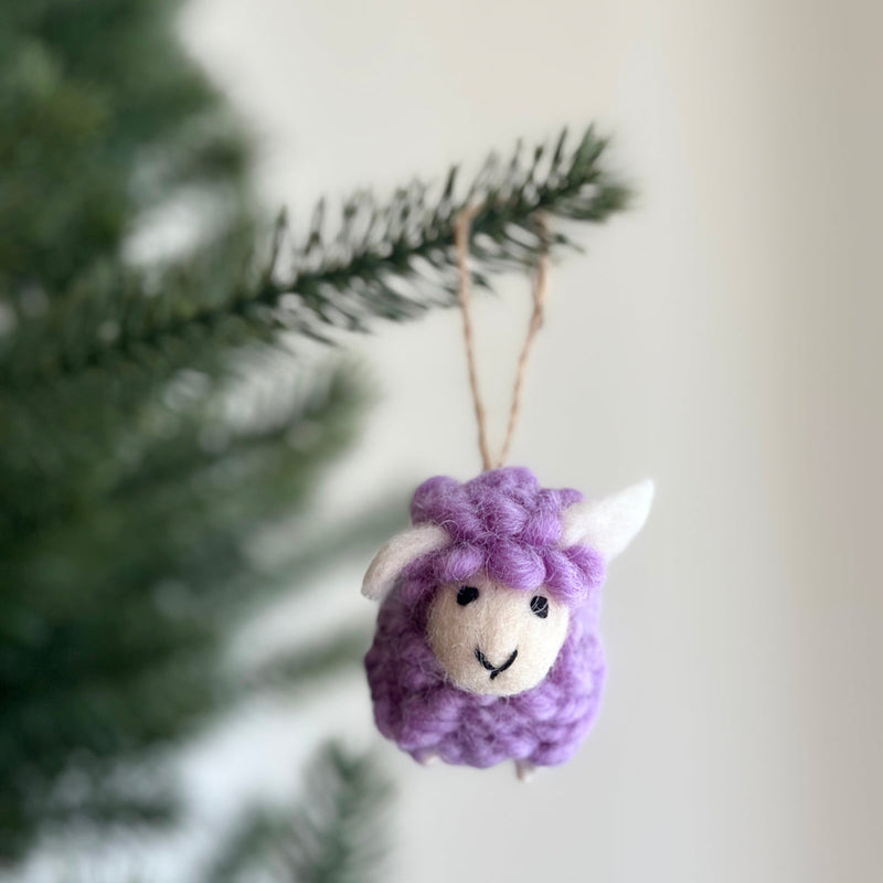 A purple felt mini fluffy sheep ornament hangs from a dark green Christmas tree branch. This small, purple wool felt sheep ornament has a fluffy, textured body, a smiley face, and small, upright white ears. A jute string is visible at the top.