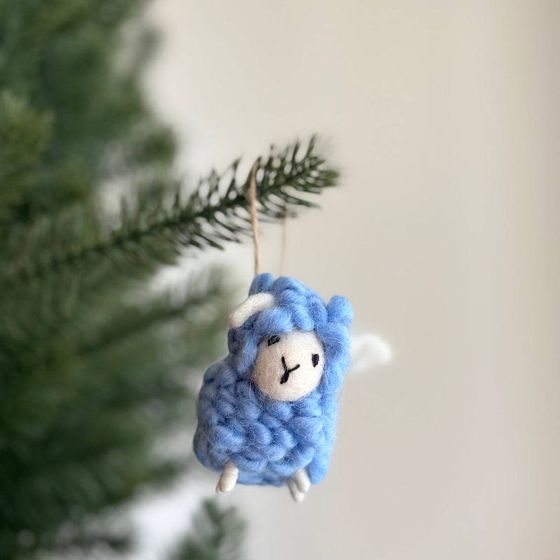 A blue wool felt mini fluffy sheep ornament hangs playfully from a Christmas tree against a soft, natural background. This handcrafted felt ornament features a fluffy woolly texture, a small round face with a stitched 'Y' marking, tiny white felt ears, and a tail. A jute string is attached at the top for hanging.