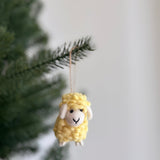 A cheerful yellow wool felt mini fluffy sheep ornament hangs from a Christmas tree branch. This delightful felted sheep features a fluffy texture with small white details for its face and feet, suspended by a jute string, ready to add a whimsical touch to holiday decor.