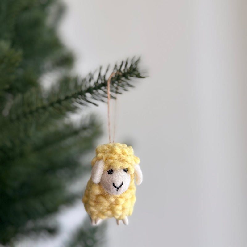 A cheerful yellow wool felt mini fluffy sheep ornament hangs from a Christmas tree branch. This delightful felted sheep features a fluffy texture with small white details for its face and feet, suspended by a jute string, ready to add a whimsical touch to holiday decor.