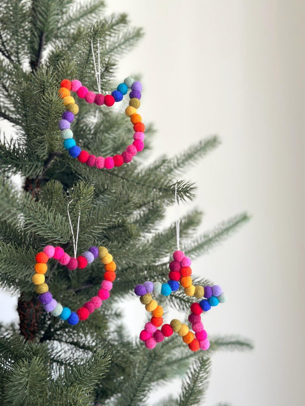 A vibrant Felt Mini Pompom Love Wreath Ornament is prominently displayed, hanging on a lush green Christmas tree. These 4-inch diameter ornaments are made from colorful 100% wool felt mini pom poms arranged in a joyful way. Suspended by a delicate jute thread, each adds a festive and handmade touch to the holiday decor.