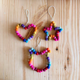 Three colorful handmade felt ornaments are arranged on a light-colored wooden surface in an overhead shot. At the upper left is a heart shape, to its right is a five-pointed star, and below them is a stylized cat head with pointed ears. Each ornament is formed from a continuous string of small, round, rainbow-colored felt pom-poms and includes a white cotton string loop for hanging. The bright colors and soft texture against the natural wood give the photo a warm, whimsical, and cheerful handmade feel.