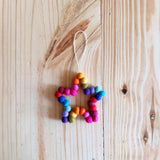 A vibrant, multi-colored felt pompom star wreath ornament rests on a light wooden surface. This 4-inch, needle-felted Christmas star ornament features cheerful, rainbow-effect wool felt pompoms, with a white jute thread attached for hanging.