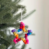 A needle felted star ornament with colorful mini pompom balls hangs from a lush Christmas tree branch, creating a joyful and festive atmosphere. This handcrafted felt ornament shaped like a Christmas star, adorned with small, vibrant rainbow pompoms. A jute thread is attached to the top, giving the felted ornament a playful, textured look as a cheerful addition to the holiday decor.